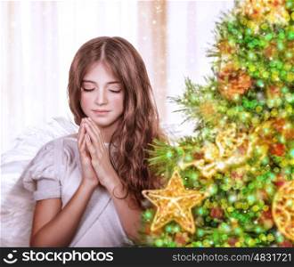 Portrait of little teen angel praying with closed eyes, standing near beautiful decorated Christmas tree at home, waiting for a miracle