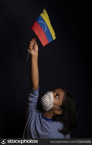 Portrait of little Venezuelan girl wearing medical mask and holding hopefully the flag of Venezuela