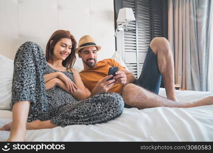 Portrait of lovely couple relaxing and using mobile phone while laying on bed at hotel room. Lifestyle and travel concept.