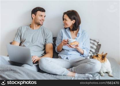 Portrait of lovely couple sit together on bed, use laptop computer, have pleasant conversation between each other, do shopping online. Jack russell terrier lies near hosts in bedroom