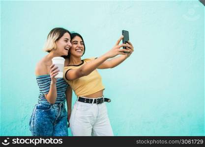 Portrait of lovely lesbian couple having fun and taking a selfie with mobile phone against light blue background. LGBT concept.