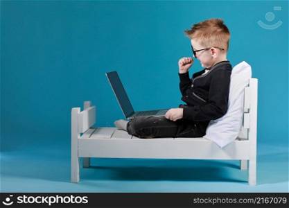 Portrait of lovely male baby lying on bedroom with laptop while wearing a round glasses and looking at camera.. Portrait of lovely male baby lying on bedroom with laptop while wearing a round glasses and looking at camera