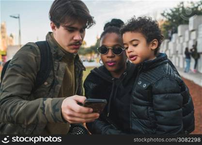 Portrait of lovely mixed race ethnic family having fun, relaxing and using mobile phone at the park outdoors.