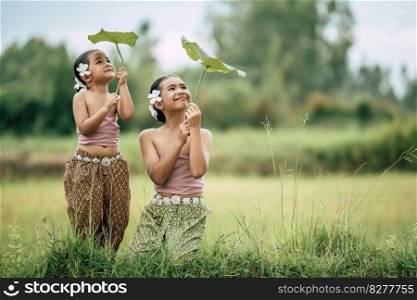 Portrait of lovely sister and young sister in Thai traditional dress and put white flower on her ear, looking to lotus’ leaf in hand and smile with happy on rice field, copy space