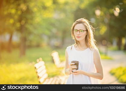 Portrait of lovely urban girl with paper cup in her hands. Happy smiling woman walking in a city park. Fashionable blonde girl wearing spectacles
