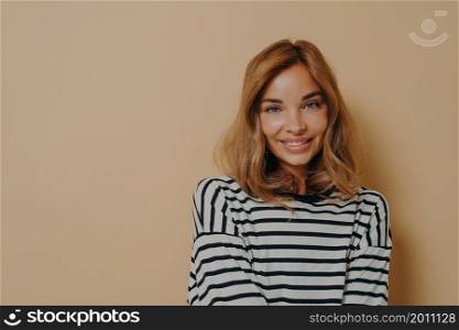 Portrait of lovely woman with minimal natural makeup smiles gently looks directly at camera wears casual striped jumper isolated over beige background copy space for your promotional content. Portrait of lovely woman with minimal natural makeup smiles gently looks directly at camera
