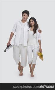 Portrait of loving young couple holding slippers while walking over white background
