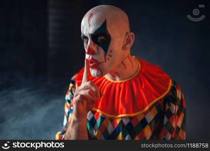 Portrait of mad bloody clown shows the quiet sign, face in blood. Man with makeup in halloween costume, maniac. Portrait of mad bloody clown shows the quiet sign