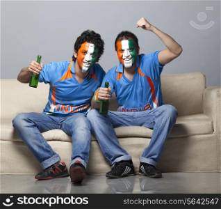 Portrait of male friends on sofa with face painted in Indian tricolor cheering while having drink at home