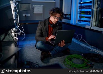 Portrait of male system administrator working in server room using laptop while inspecting in computer. Database management and maintenance concept. A system administrator working in server room using laptop