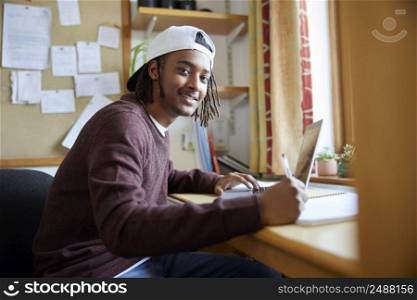 Portrait Of Male University Or College Student Wearing Baseball Cap Studying With Laptop At Desk In Room