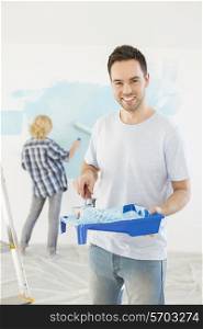 Portrait of man holding paint roller and tray with woman painting wall in background