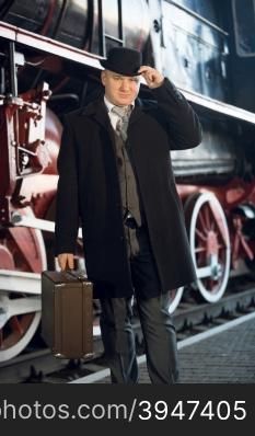 Portrait of man in retro suit, bowler hat and suitcase posing at old locomotive