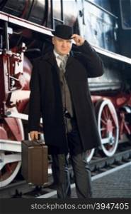 Portrait of man in retro suit, bowler hat and suitcase posing at old locomotive