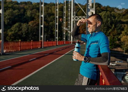 Portrait of man runner taking break from outdoor jogging. Sportsman leaning on bridge railing and relaxing after running. Portrait of man runner taking break from outdoor jogging