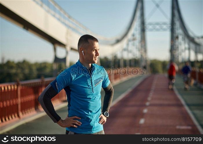 Portrait of man runner taking break from outdoor jogging. Sportsman leaning on bridge railing and relaxing after running. Portrait of man runner taking break from outdoor jogging