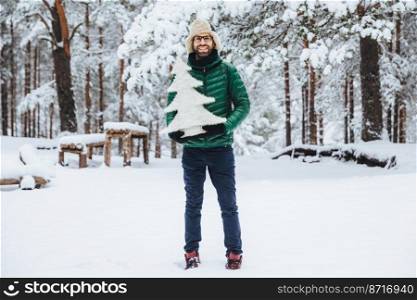 Portrait of man with thick beard and musatche has pleased expression, walks in beautiful winter forest, holds small white artificial fir tree, advertises ir before coming New Year holidays