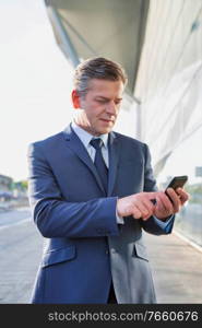 Portrait of mature attractive businessman using smartphone while standing in front of airport