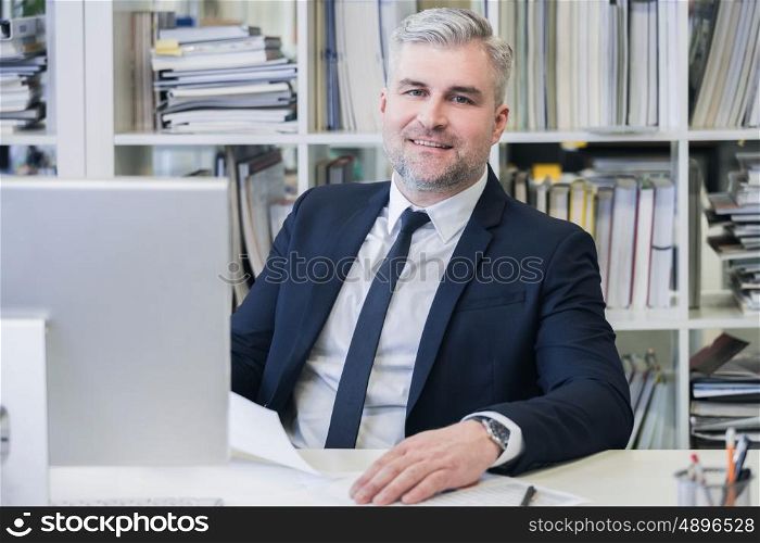 Portrait of mature business man. Portrait of mature business man with grey hair in his modern office with computer and many file folders on shelves on background