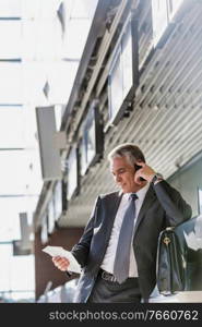 Portrait of mature businessman talking on smartphone while looking at his boarding pass in airport