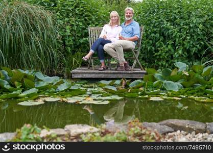 Portrait Of Mature Couple Celebrating With Champagne Sitting On Chairs On Wooden Jetty By Lake