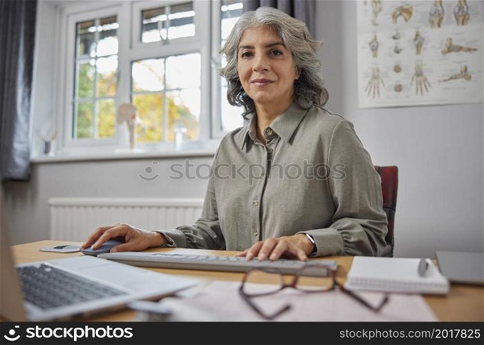 Portrait Of Mature Female GP At Desk In Doctors Office