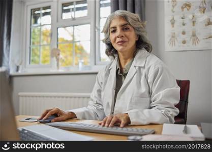 Portrait Of Mature Female GP Wearing White Coat At Desk In Doctors Office
