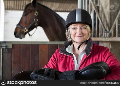Portrait Of Mature Female Owner Holding Saddle In Stable With Horse