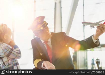 Portrait of mature pilot holding airplane toy while playing with cute little girl in airport