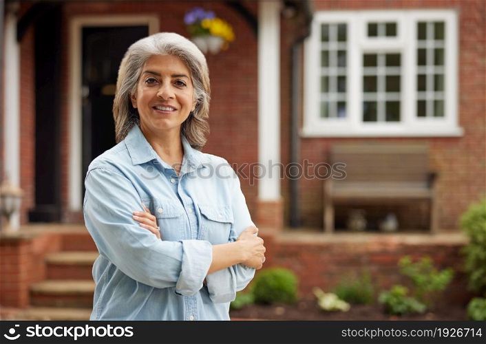 Portrait Of Mature Woman Standing In Garden In Front Of Dream Home In Countryside
