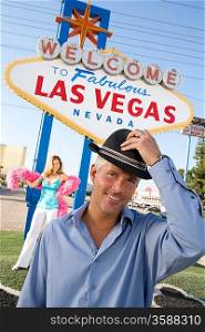 Portrait of mid-adult man in front of Welcome to Las Vegas sign, mid-adult woman in background.
