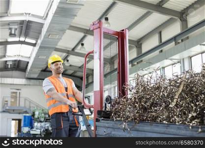 Portrait of mid adult worker pulling hand truck loaded with steel shavings in factory