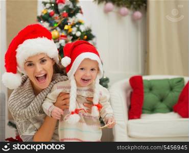 Portrait of mom and eat smeared baby girl in Christmas hats near Christmas tree