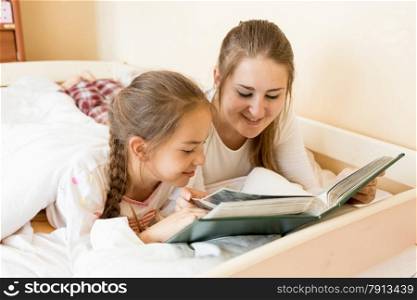 Portrait of mother reading big old book to daughter at bed
