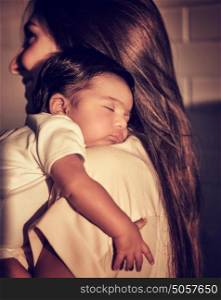 Portrait of mother standing back side and caring little sleeping baby on shoulders, enjoying mild sunset light, happy young family, peace concept
