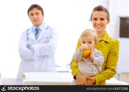 Portrait of mother with baby holding apple and doctor in background&#xA;