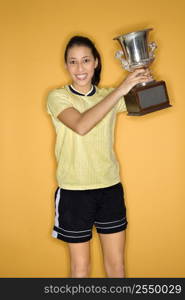 Portrait of Multi-racial teen girl in soccer uniform holding trophy and smiling standing against yellow background.