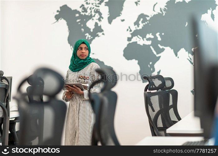 Portrait of Muslim female software developer with green hijab holding tablet computer while standing at modern open plan startup office. High-quality photo. Portrait of muslim female software developer with green hijab holding tablet computer while standing at modern open plan startup office