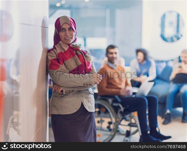 Portrait of Muslim woman wearing hijab in a modern open space coworking office. Middle eastern Successful Arab businesswoman in creative startup office. Disabled businessman in a wheelchair on team meeting in background.. Portrait of young muslim woman wearing hijab in office while looking at camera. Close up face of arabic business woman covered with headscarf smiling. Successful arab businesswoman in modern office.