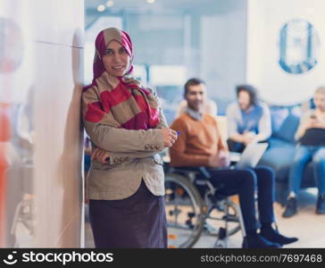 Portrait of Muslim woman wearing hijab in a modern open space coworking office. Middle eastern Successful Arab businesswoman in creative startup office. Disabled businessman in a wheelchair on team meeting in background.. Portrait of young muslim woman wearing hijab in office while looking at camera. Close up face of arabic business woman covered with headscarf smiling. Successful arab businesswoman in modern office.