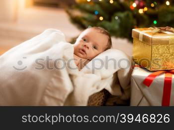 Portrait of newborn baby boy lying under blanket next to Christmas tree and gift boxes