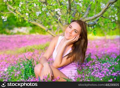Portrait of nice dreamy girl sitting down on pink flowers field in apple tree garden, watching for first spring blossom, beauty of nature concept
