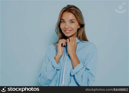 Portrait of nice looking female with long brunette hair. Folding hands at neck and holding blouse collar. Wearing blue shirt with long sleeves. Smiling at camera isolated over pastel blue background. Portrait of attractive, nice looking female with long brunette hair