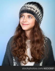 Portrait of nice stylish teen model wearing warm hat and posing over blue background, dreamy looking in side, winter fashion for girls