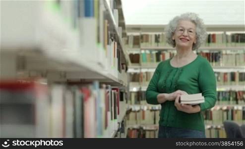 Portrait of old retired woman choosing book in library and taking it from shelf. Dolly shot