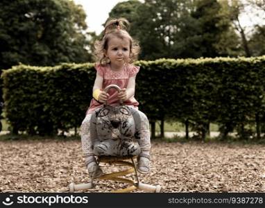 Portrait of one beautiful caucasian baby girl riding on a wooden spring swing with a potato chip in her mouth at the playground in the city park, close-up side view.. Portrait of a beautiful caucasian girl on a swing in the park.