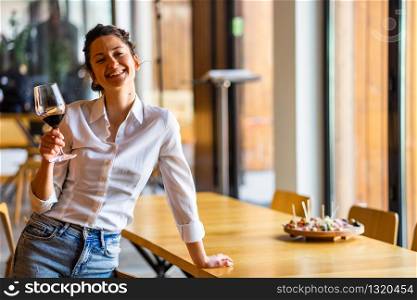 Portrait of one young beautiful caucasian woman standing by the table leaning holding glass of red wine at home alone smiling wearing white shirt looking to the camera front view