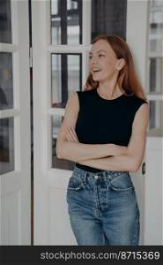Portrait of overjoyed young caucasian woman wearing casual clothes on white door background laugh at funny joke. Smiling female feels optimistic, standing with crossed arms at home.. Smiling young caucasian woman in casual clothes on white door background laugh at funny joke at home