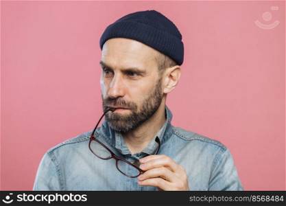 Portrait of pensive bearded man takes off glasses, wears denim stylish jacket and hat, isolated over pink background. Middle aged unshaven male contemplates about something, looks concentrated