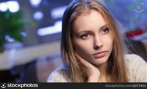 Portrait of pensive charming girl with amazing blonde long hair and grey eyes sitting in cafe with her hand on chin, remembering unforgettable moments and smiling at camera with perfect toothy smile.